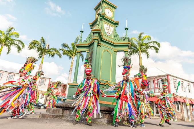 Masquerade dancers in bright ribboned costumes and peacock-feather headdresses at the Berkeley Memorial clock, the Circus, Basseterre